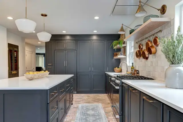 Kitchen by ELM Construction with charcoal cabinetry, white counters, a brick floor and copper pans on open shelves